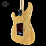 The back of a natural wood electric guitar, showing the grain, body contour, pickguard, and hardware against a black background.
