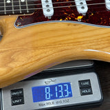 Close-up of a guitar body resting on a digital scale, showing the wood grain finish and part of the headstock area. The scale displays 8.