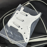 A white guitar pickguard with pre-drilled mounting holes, sealed in a clear plastic bag and resting on a black guitar case.