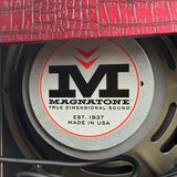 Close-up of a round Magnatone speaker with a silver metallic center label featuring a large black 'M' logo and red accents, with part of a textured red surface visible above.