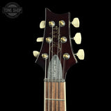 Close-up of the headstock of a McCurdy 334 acoustic guitar against a solid black background, showing the dark reddish-brown wood, gold tuning pegs, and cream-colored tuners.