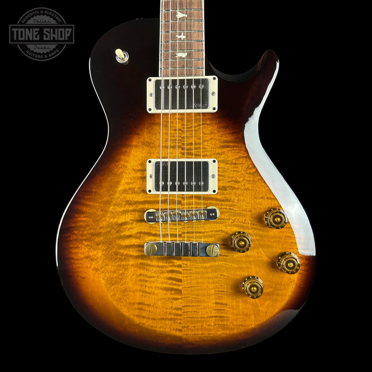 A close-up of an electric guitar body showing a vibrant sunburst finish that transitions from a bright amber-orange center to a dark brown edge. The guitar features gold hardware, dual humbucker pickups, and a dark fretboard, photographed against a solid black background with a Tone Shop watermark in the top-left corner.