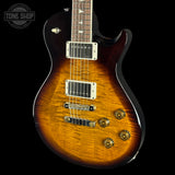 A close-up of an electric guitar body showing a sunburst flame finish that transitions from bright amber-orange in the center to dark brown and black at the edges. The guitar features gold hardware, white pickups, and a dark fretboard, photographed against a solid black background with a Tone Shop watermark in the top-left corner.