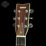 Close-up of a Yamaha acoustic guitar headstock with a dark wood finish, gold lettering, and silver tuning keys against a black background.