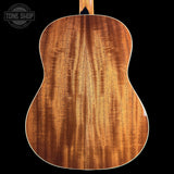 Close-up of the back of an acoustic guitar showing a rich, figured wood grain finish in warm brown tones, set against a solid black background with a Tone Shop watermark in the top left corner.