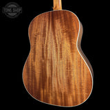 Close-up of the back of an acoustic guitar showing a rich, warm brown wood grain finish with white binding, set against a solid black background. A Tone Shop watermark is visible in the top-left corner.