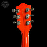 Close-up of the headstock of a guitar with a vibrant orange-red finish and six silver tuning pegs, set against a solid black background. A Tone Shop watermark is visible in the top left corner.