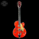A Gretsch electric guitar with a vibrant orange-red cherry finish, shown against a solid black background. The guitar features a cream pickguard, chrome hardware, and a dark fretboard.