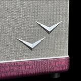Close-up of a vintage-style speaker cabinet featuring a beige woven fabric grille, two silver V-shaped metal badges, and a pink crocodile-embossed lower trim against a black background.