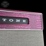 Close-up of a guitar amplifier corner with a pink crocodile-embossed top panel, a black Tone logo badge, and a gray fabric grille below on a black background.