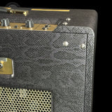 Close-up of a guitar amplifier cabinet corner showing textured black and gray fabric-covered edges, gold metal trim, control knobs, and a perforated speaker grille against a black background.