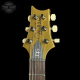 Close-up of the headstock of a gold-finish electric guitar against a black background, showing six cream-colored tuning pegs, a dark wood fretboard, and a Tone Shop watermark in the upper left corner.
