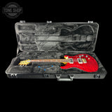 A red electric guitar with a yellow headstock resting inside an open black hard case with gray foam lining, shown against a black background. A Tone Shop watermark is visible in the top-left corner.