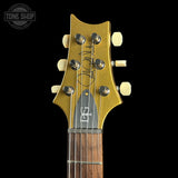Close-up of the headstock of a gold-finish electric guitar against a black background, with a Tone Shop watermark in the upper left corner.
