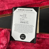 A Gibson Custom Certificate of Authenticity card inserted between two black cases, placed on a plush red fabric background. The certificate includes handwritten details and a Gibson logo seal at the bottom.