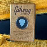 Gibson Custom guitar pick in a wooden case with engraved text on a textured yellow background.