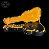 A black acoustic guitar resting inside an open black hard case with a bright yellow-orange velvet interior, shown against a solid black background. A Tone Shop watermark is visible in the top-left corner.