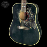 A close-up of a dark acoustic guitar body with gold decorative inlay around the soundhole and pickguard, shown against a black background. A Tone Shop watermark is visible in the upper left corner.