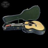 An acoustic guitar resting inside an open hard case on a black background. The guitar has a natural wood top and a dark fretboard, while the case is dark blue-green with a green interior.