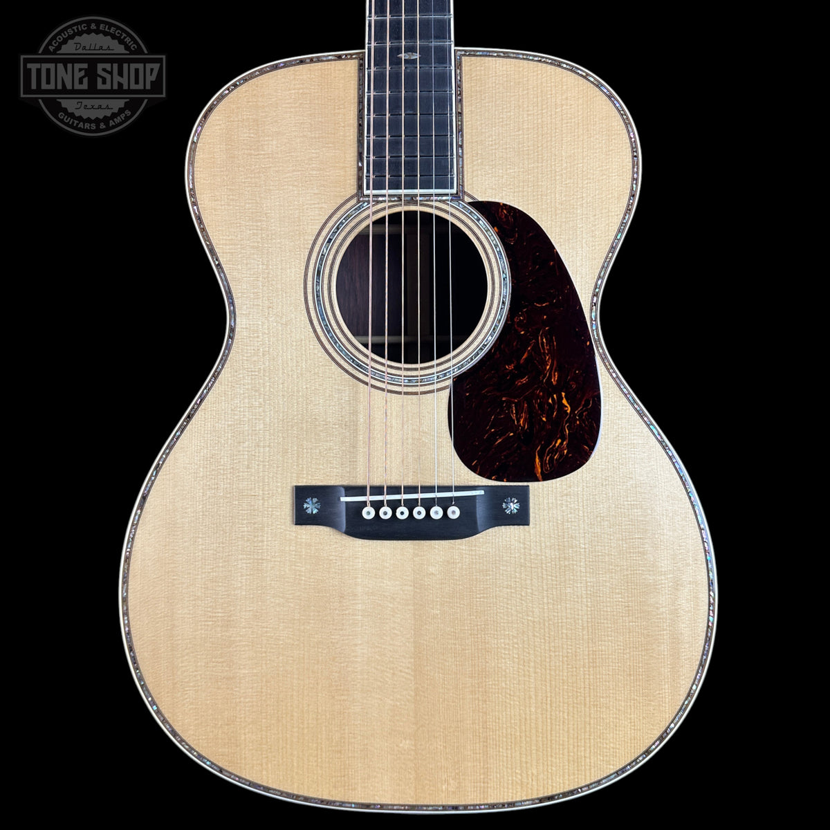 Front view of an acoustic guitar with a natural wood finish, black pickguard, and dark fretboard, shown against a solid black background with a Tone Shop watermark in the top left corner.