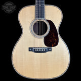Front view of an acoustic guitar with a natural wood finish, black pickguard, and dark fretboard, shown against a solid black background with a Tone Shop watermark in the top left corner.