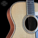 Close-up of an acoustic guitar body showing the natural wood grain, sound hole, rosette, and part of the dark fretboard against a black background. A Tone Shop watermark is visible in the top-left corner.