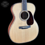 An acoustic guitar with a natural wood top, dark brown sides, and a dark tortoiseshell pickguard, shown against a solid black background with a Tone Shop watermark in the upper left.