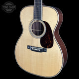 A close-up of a wooden acoustic guitar body with a natural light wood top, dark brown sides, a black pickguard, and a decorative rosette around the sound hole, shown against a black background with a Tone Shop watermark in the upper left.