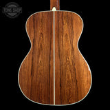 The back of an acoustic guitar made of rich brown wood with a central light-colored binding and a vertical grain pattern, shown against a solid black background with a Tone Shop watermark in the top left.