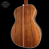 The back of an acoustic guitar made of rich, warm brown wood with a white binding edge, shown against a solid black background. A Tone Shop watermark is visible in the top-left corner.