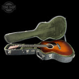 An acoustic guitar with a sunburst finish displayed inside an open hard case on a solid black background. The case is olive green velvet-lined with a gray exterior.
