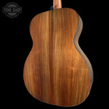Close-up of the back of an acoustic guitar showing a rich, warm brown wood grain finish, set against a solid black background with a Tone Shop watermark in the top left corner.
