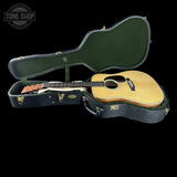 An acoustic guitar with a natural wood top and dark neck, resting inside an open black hard case with a green felt lining, shown against a solid black background. A Tone Shop watermark is visible in the top-left corner.