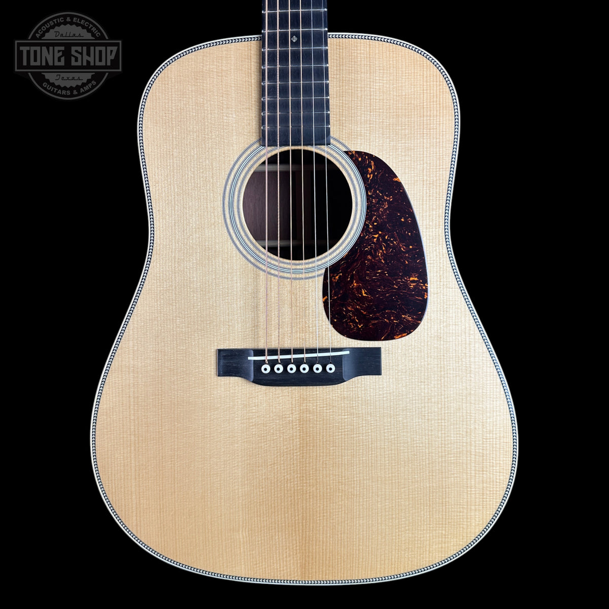 A front-facing acoustic guitar with a natural wood top, dark fretboard, and dark tortoiseshell-style pickguard, shown against a solid black background. A Tone Shop logo is visible in the upper left corner.
