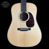 A front-facing acoustic guitar with a natural wood top, dark fretboard, and dark tortoiseshell-style pickguard, shown against a solid black background. A Tone Shop logo is visible in the upper left corner.