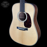 A close-up studio shot of an acoustic guitar against a solid black background, featuring a light natural wood top, dark brown sides, and a glossy tortoiseshell pickguard. A Tone Shop watermark is visible in the upper left corner.