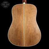 The back of an acoustic guitar made of medium brown wood with a light-colored center stripe and white binding, shown against a solid black background. A Tone Shop watermark is visible in the top left corner.