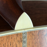 Close-up of a guitar body corner with a cream-colored triangular heel pad resting on the edge, showing dark brown wood and a decorative inlay.