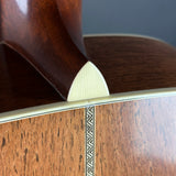 Close-up detail of an acoustic guitar body showing the dark brown wood back, a cream-colored heel, a gold-toned edge strip, and a decorative inlay on the front.