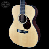A close-up studio shot of an acoustic guitar body against a solid black background, featuring a natural wood grain top, dark sides, and a black pickguard. A Tone Shop logo is visible in the top-left corner.