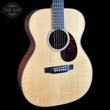 An acoustic guitar with a natural wood top and dark reddish-brown sides, shown against a solid black background. A Tone Shop logo is visible in the top-left corner.