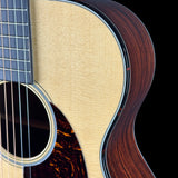 Close-up detail of an acoustic guitar showing the natural wood body, sound hole rosette, pickguard, and part of the dark fretboard against a black background.