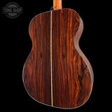 Close-up of the back of an acoustic guitar showing a rich, dark reddish-brown wood grain finish with a central gold-toned inlay, set against a solid black background.