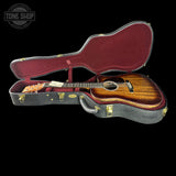 An acoustic guitar with a glossy brown wooden body resting inside an open black hard case with a burgundy interior, photographed against a solid black background. A Tone Shop watermark is visible in the top-left corner.