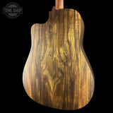 Close-up of the back of an acoustic guitar body made from richly figured brown wood, showing prominent grain patterns and a cutaway shape, set against a solid black background with a Tone Shop watermark in the top left.
