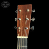 Close-up of an acoustic guitar headstock with a reddish-brown wood finish and six silver tuning pegs, shown against a solid black background. A Tone Shop watermark is visible in the top-left corner.