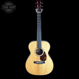 A natural wood acoustic guitar with a dark headstock and black pickguard, shown against a solid black background with a Tone Shop watermark in the top left corner.