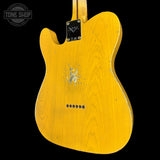 The back of a yellow-finish electric guitar, showing the wood grain, a scuffed center-pull tailpiece, and a black plate with a stylized logo, photographed against a black background with a Tone Shop watermark in the top left.