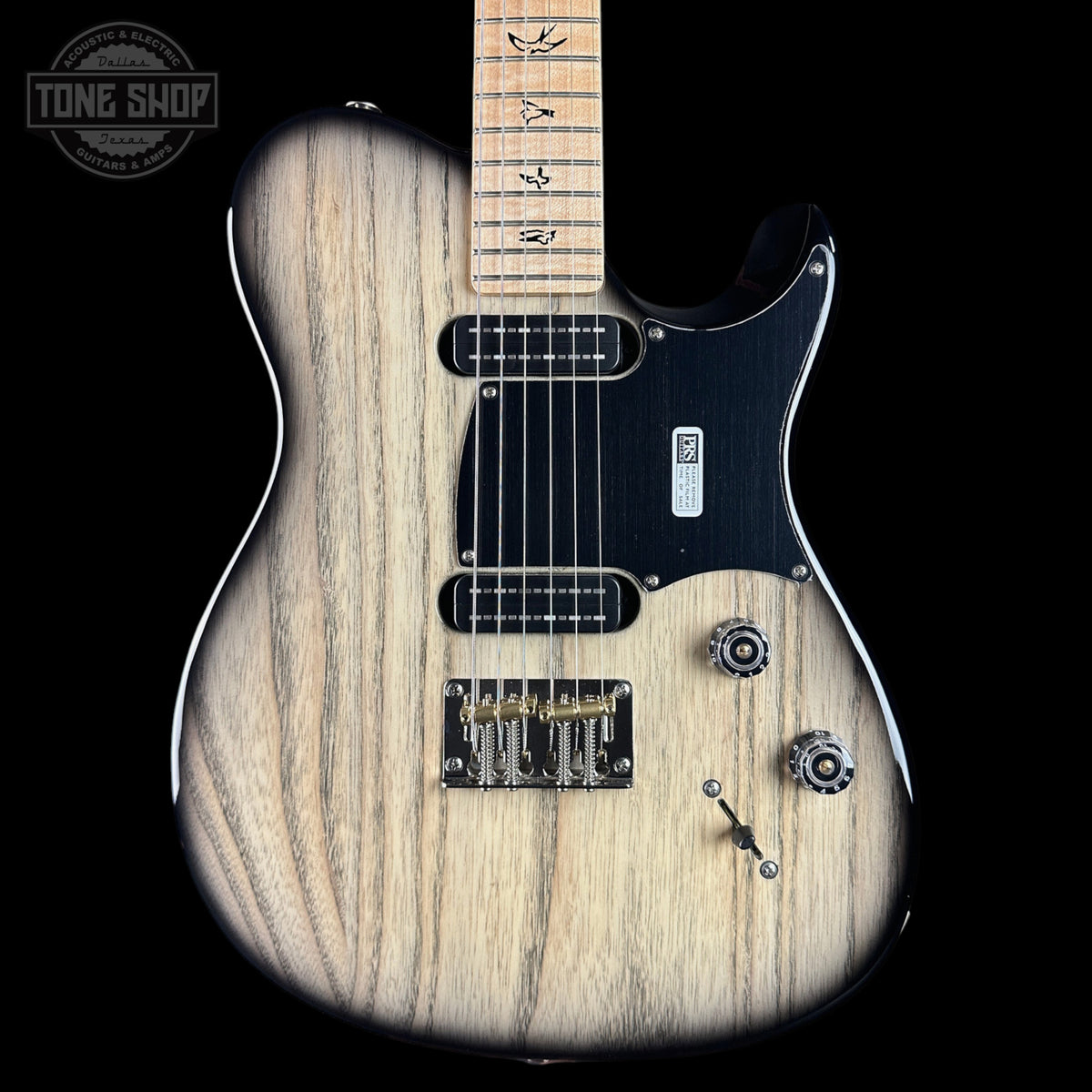 A close-up of an electric guitar body showing a natural wood grain finish, black pickguard, dual humbucker pickups, and control knobs against a black background. A Tone Shop watermark is visible in the top left corner.