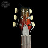Close-up of a custom electric guitar headstock featuring a sunburst finish that transitions from black to red and orange, with cream-colored tuning pegs and a silver metal 'Custom' plate on the neck. The headstock is set against a solid black background with a Tone Shop watermark in the upper left corner.